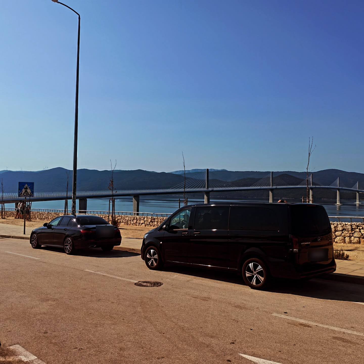 A black van and a black car parked by the sea with the Pelješac Bridge behind